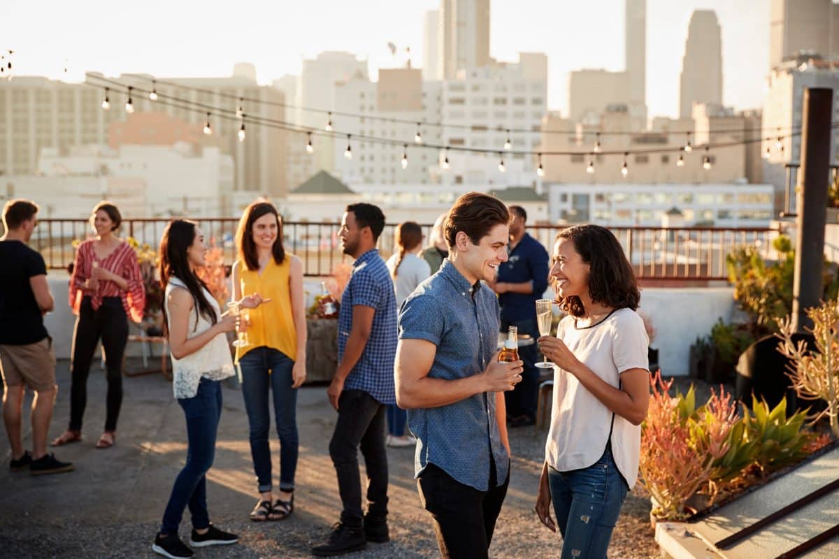 Un groupe de personnes discutent sur un toit, avec la silhouette de la ville en arrière plan. Certains boivent, d'autres discutent, et des guirlandes lumineuses sont visibles au dessus. Friends Gathered On Rooftop Terrace For Party With City Skyline In Background