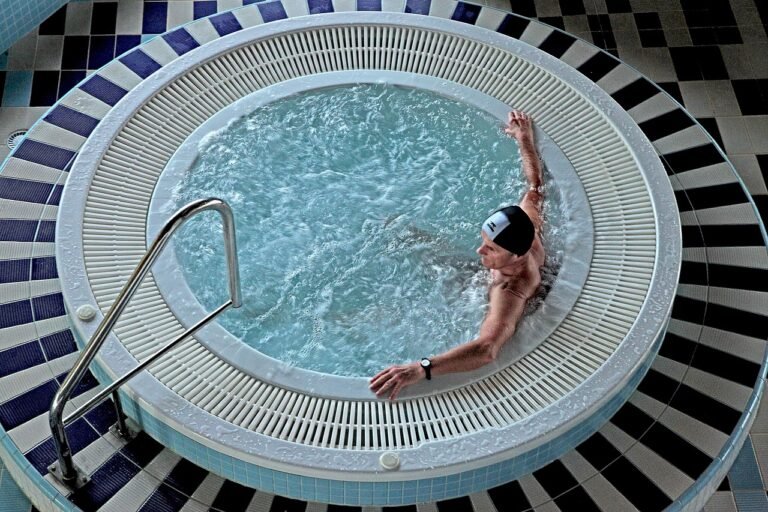 Une personne portant un bonnet de bain se détend dans un jacuzzi intérieur circulaire à l'eau bouillonnante, à côté de mains courantes en métal et d'un sol carrelé.