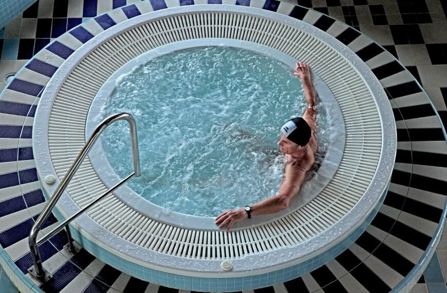 Une personne portant un bonnet de bain se détend dans un jacuzzi intérieur circulaire à l'eau bouillonnante, à côté de mains courantes en métal et d'un sol carrelé.