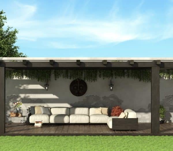 Une pergola en bois avec un canapé blanc et des coussins trône dans un jardin. Une cheminée se trouve sur la droite et l'espace est entouré de verdure.