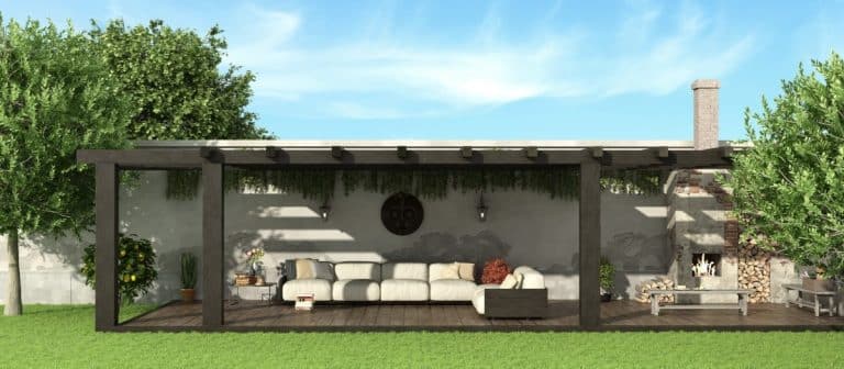 Une pergola en bois avec un canapé blanc et des coussins trône dans un jardin. Une cheminée se trouve sur la droite et l'espace est entouré de verdure.