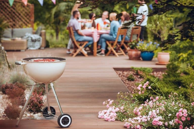 Un barbecue au charbon de bois blanc trône au premier plan sur une terrasse de jardin, avec un groupe de personnes assises autour d'une table à l'arrière plan. Des fleurs roses et des plantes en pot entourent la scène.
