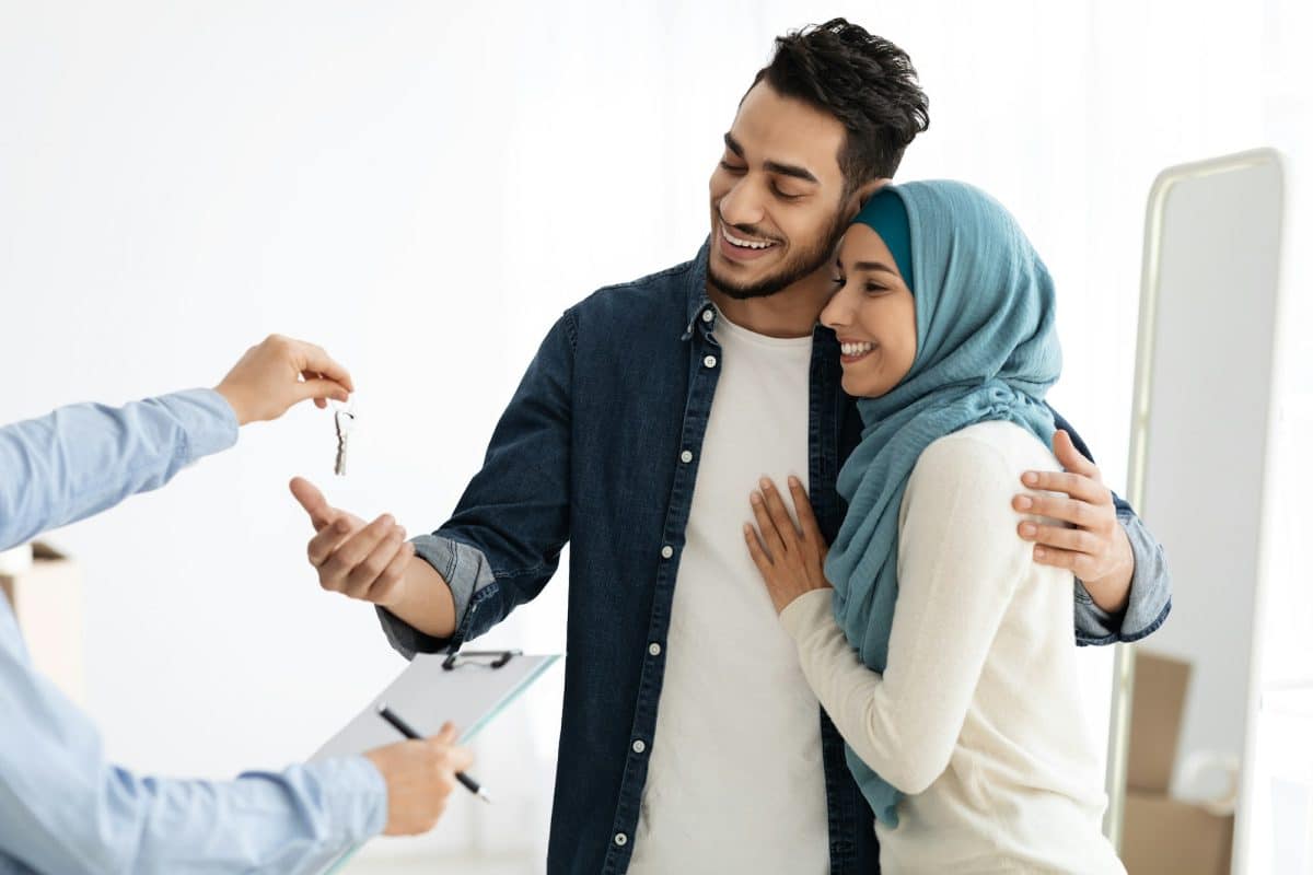 Un couple souriant reçoit les clés de la maison des mains d'une personne, ce qui signifie l'achat ou la location d'un bien immobilier. L'homme a son bras autour de la femme, qui porte un foulard sur la tête.