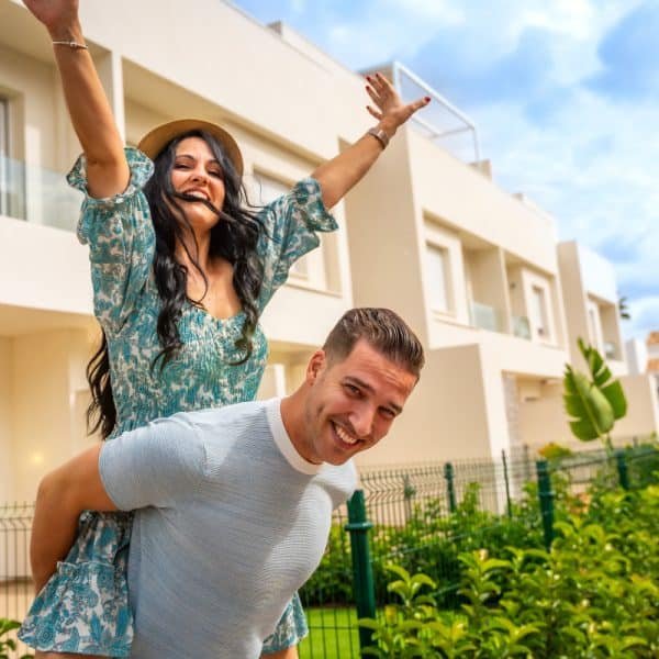 Une femme au chapeau chevauche joyeusement un homme souriant, tous deux en tenue décontractée. Ils sont devant un bâtiment moderne entouré de verdure. Le ciel est partiellement nuageux.