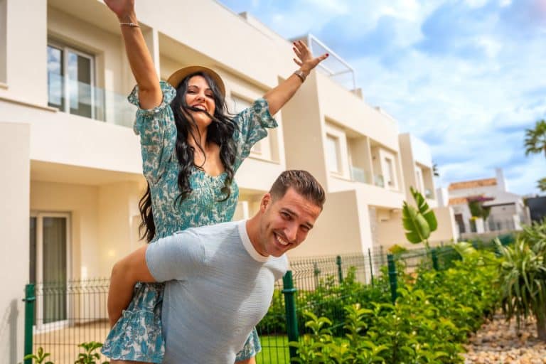 Une femme au chapeau chevauche joyeusement un homme souriant, tous deux en tenue décontractée. Ils sont devant un bâtiment moderne entouré de verdure. Le ciel est partiellement nuageux.