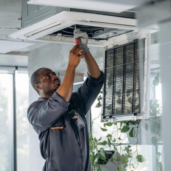 Un technicien en uniforme gris répare un climatiseur monté au plafond à l'aide d'un tournevis dans une pièce moderne avec des plantes.