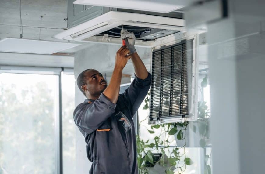 Un technicien en uniforme gris répare un climatiseur monté au plafond à l'aide d'un tournevis dans une pièce moderne avec des plantes.