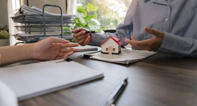 Deux personnes à une table discutent de documents. L'une d'elles pointe du doigt une petite maquette de maison avec un stylo, suggérant une discussion sur l'immobilier ou la propriété. Des papiers et une pile de dossiers sont visibles.