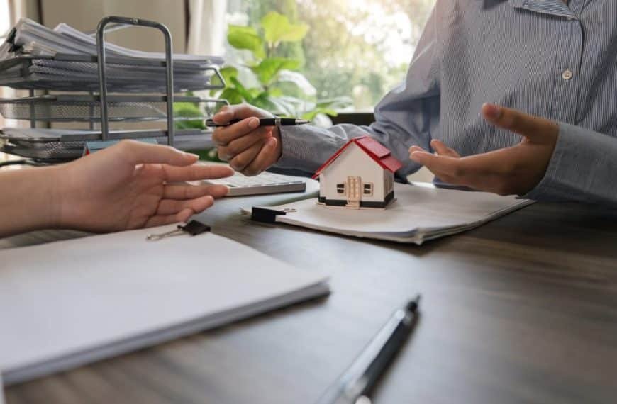 Deux personnes à une table discutent de documents. L'une d'elles pointe du doigt une petite maquette de maison avec un stylo, suggérant une discussion sur l'immobilier ou la propriété. Des papiers et une pile de dossiers sont visibles.