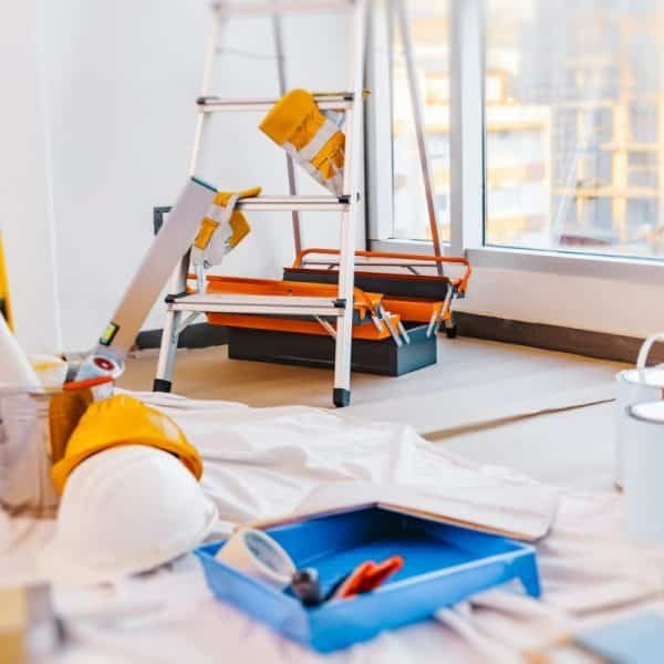 Pièce en rénovation avec une échelle, des pots de peinture, des rouleaux de papier, des outils et un casque de sécurité sur une toile de protection près d'une grande fenêtre.