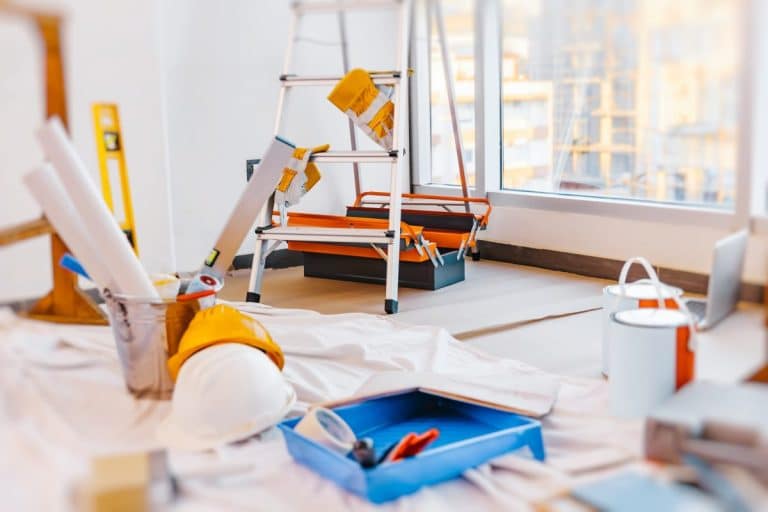 Pièce en rénovation avec une échelle, des pots de peinture, des rouleaux de papier, des outils et un casque de sécurité sur une toile de protection près d'une grande fenêtre.
