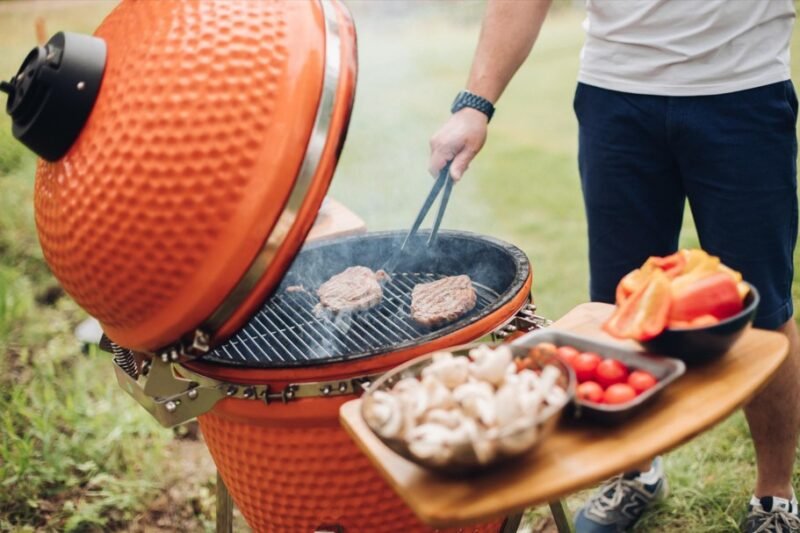 Une personne fait griller des steaks sur un barbecue en céramique orange, tandis que des légumes, des champignons et des tomates sont disposés sur un plateau en bois à proximité.