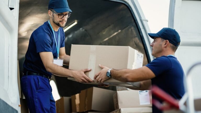 Deux travailleurs portant des uniformes bleus et des casquettes chargent des cartons dans une camionnette de livraison. L'un d'eux remet un carton à l'autre. Plusieurs cartons sont visibles à l'intérieur de la camionnette.