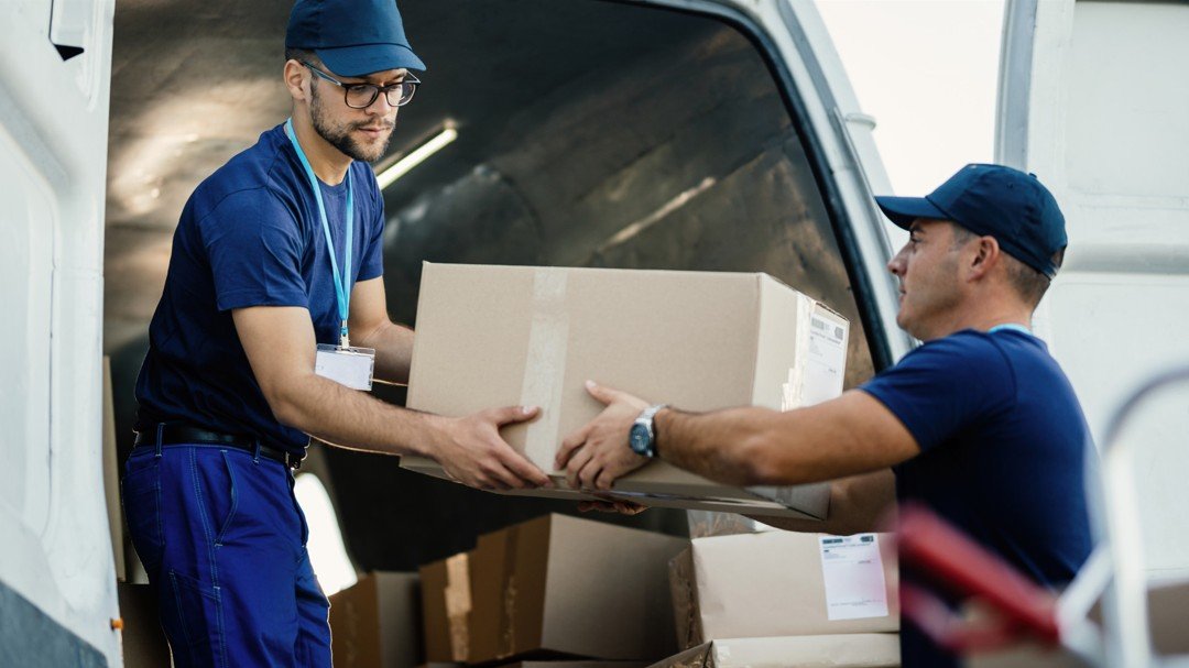 Deux travailleurs portant des uniformes bleus et des casquettes chargent des cartons dans une camionnette de livraison. L'un d'eux remet un carton à l'autre. Plusieurs cartons sont visibles à l'intérieur de la camionnette.