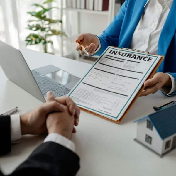 Deux personnes sont assises à un bureau et discutent d'assurance. L'une d'elles, vêtue d'un blazer bleu, tient un formulaire d'assurance et une clé. L'autre personne a les mains jointes. Un ordinateur portable et une maquette de maison sont posés sur le bureau.