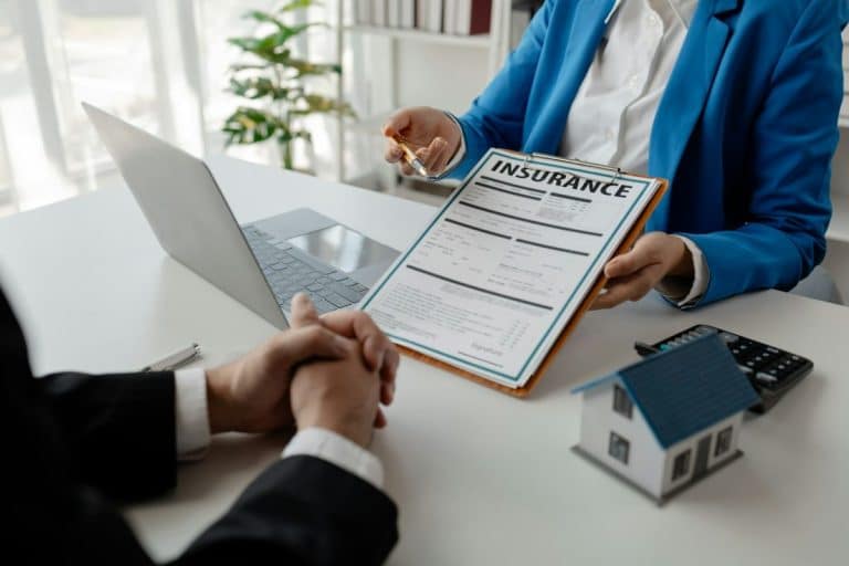 Deux personnes sont assises à un bureau et discutent d'assurance. L'une d'elles, vêtue d'un blazer bleu, tient un formulaire d'assurance et une clé. L'autre personne a les mains jointes. Un ordinateur portable et une maquette de maison sont posés sur le bureau.
