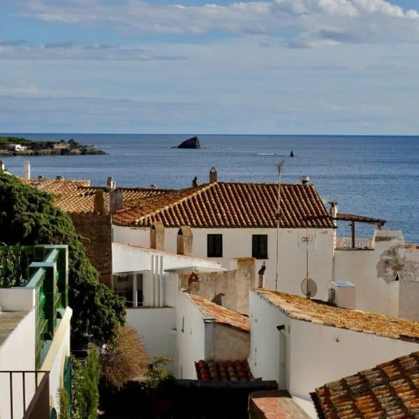 Un village côtier aux maisons blanches et aux toits en terre cuite surplombe la mer bleue calme sous un ciel partiellement nuageux.
