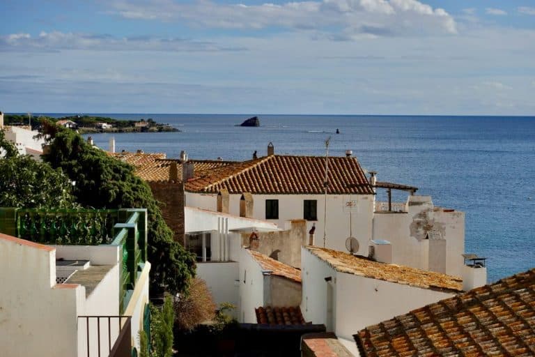 Un village côtier aux maisons blanches et aux toits en terre cuite surplombe la mer bleue calme sous un ciel partiellement nuageux.