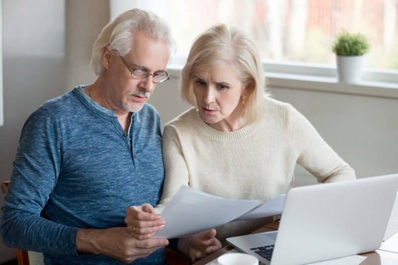 Un couple âgé consulte des papiers sur un ordinateur portable.