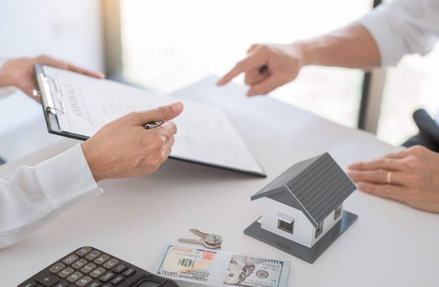 Deux individus discutant d'une transaction immobilière avec un contrat, un modèle de maison, une calculatrice et une devise sur la table.