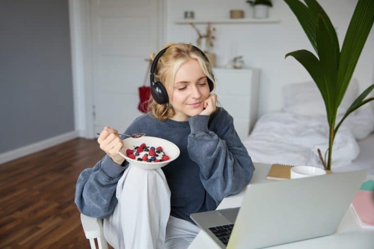 Une jeune femme portant des écouteurs est assise à un bureau avec un ordinateur portable, mangeant un bol de yaourt et des baies dans une chambre à coucher moderne et lumineuse.