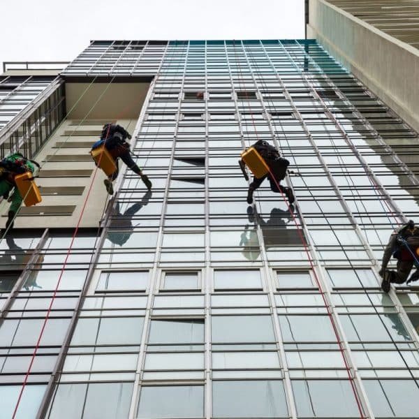 Des ouvriers sur des plates formes suspendues nettoient les fenêtres d'un grand bâtiment moderne en verre, à l'aide de cordes et d'équipements.