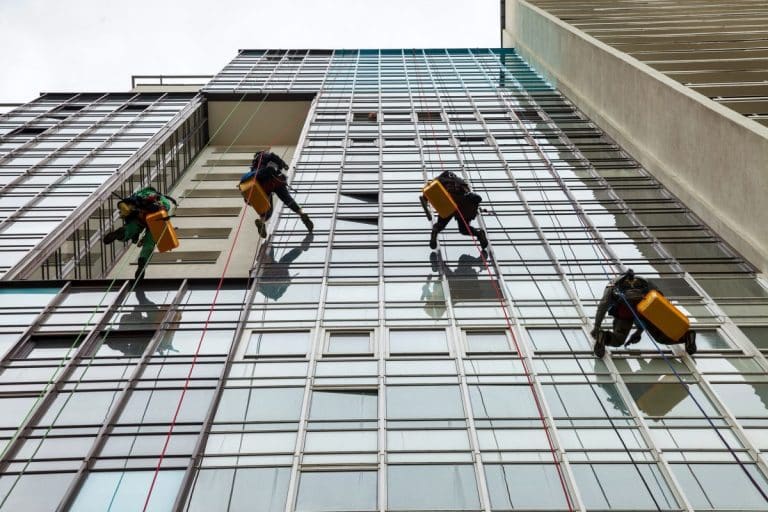 Des ouvriers sur des plates formes suspendues nettoient les fenêtres d'un grand bâtiment moderne en verre, à l'aide de cordes et d'équipements.