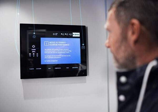 Un homme regarde un écran numérique affichant un avis de maintenance de l'ascenseur et d'autres informations dans un bâtiment moderne.