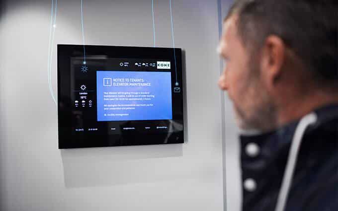 Un homme regarde un écran numérique affichant un avis de maintenance de l'ascenseur et d'autres informations dans un bâtiment moderne.