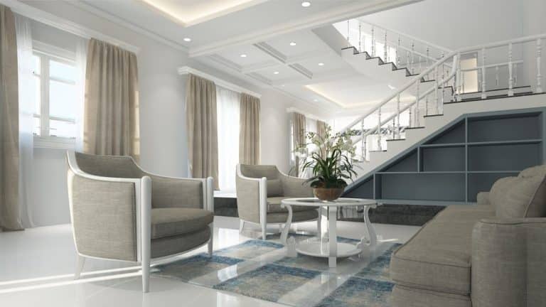 Salon moderne avec des meubles gris clair, une table basse ronde, un tapis d'accent bleu, de grandes fenêtres avec des rideaux beiges et un escalier avec des étagères encastrées.
