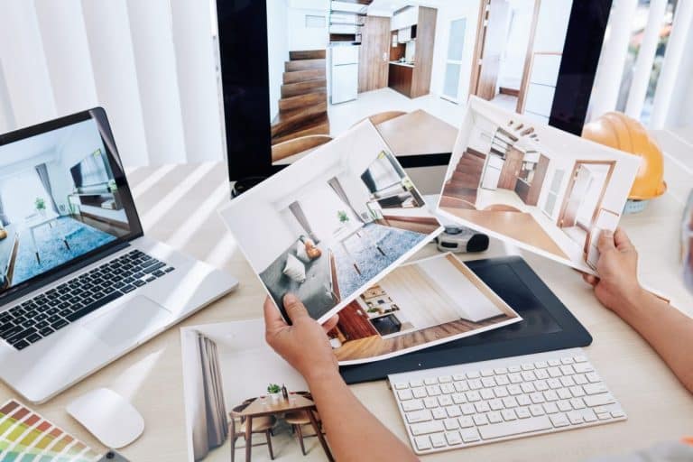 Personne tenant des photos imprimées de décorations d'intérieur, assise à un bureau avec un ordinateur portable, un écran et un clavier.