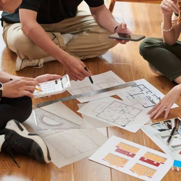 Trois personnes assises par terre discutent de plans architecturaux et d'échantillons de couleurs, à l'aide de calculatrices et de smartphones.