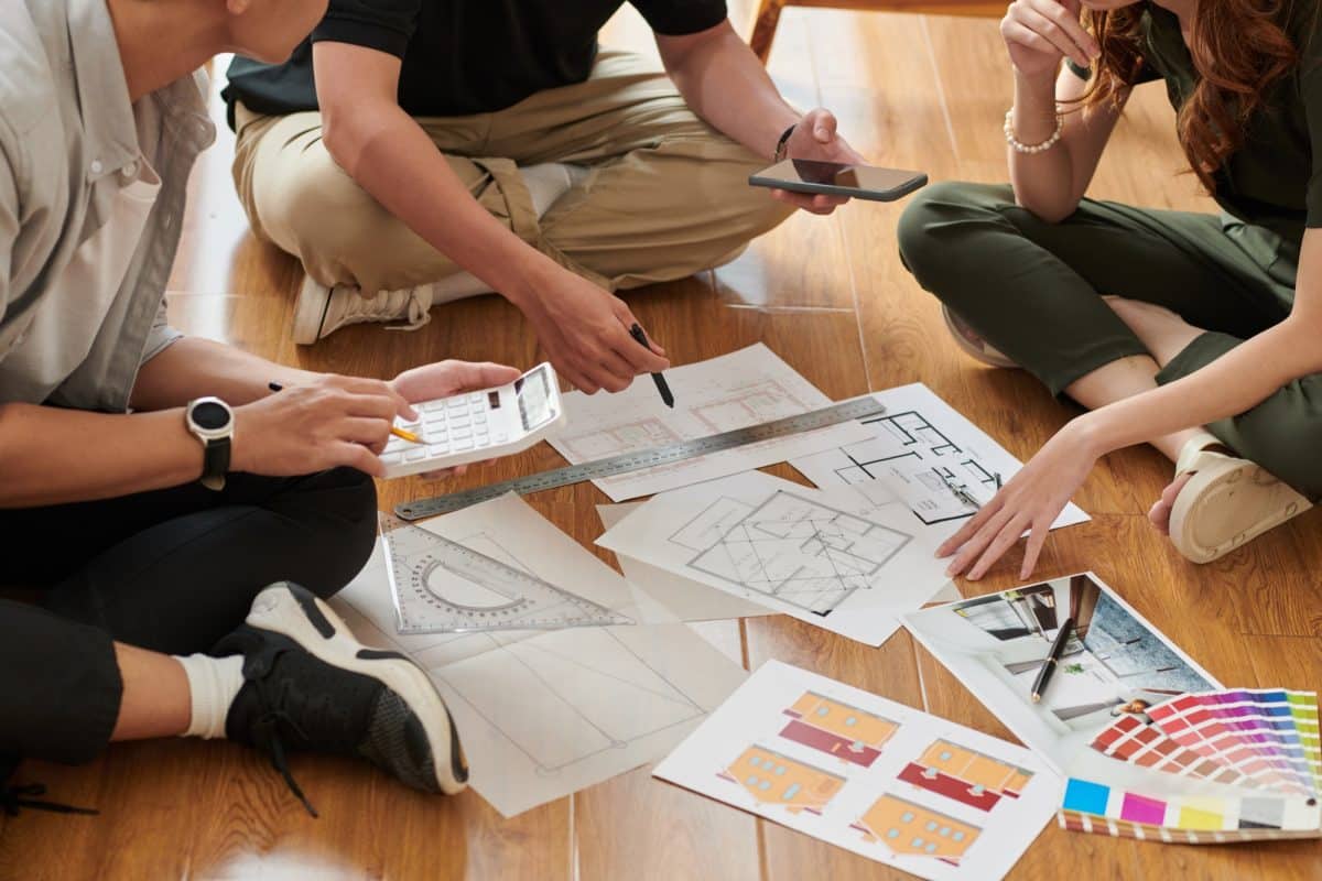 Trois personnes assises par terre discutent de plans architecturaux et d'échantillons de couleurs, à l'aide de calculatrices et de smartphones.
