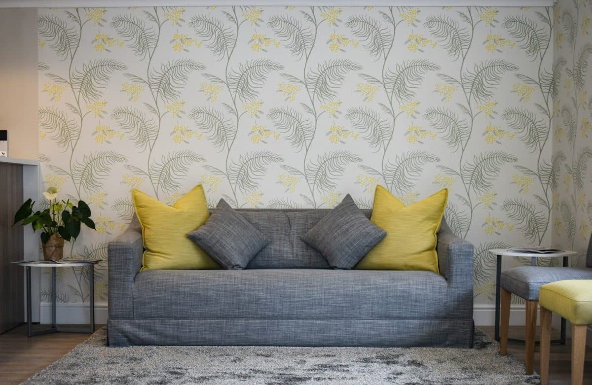 Un canapé convertible gris avec des coussins jaunes et gris est posé contre un mur tapissé d'un motif floral. Deux petites tables avec des plantes en pot et des tabourets sont placées de chaque côté du canapé.