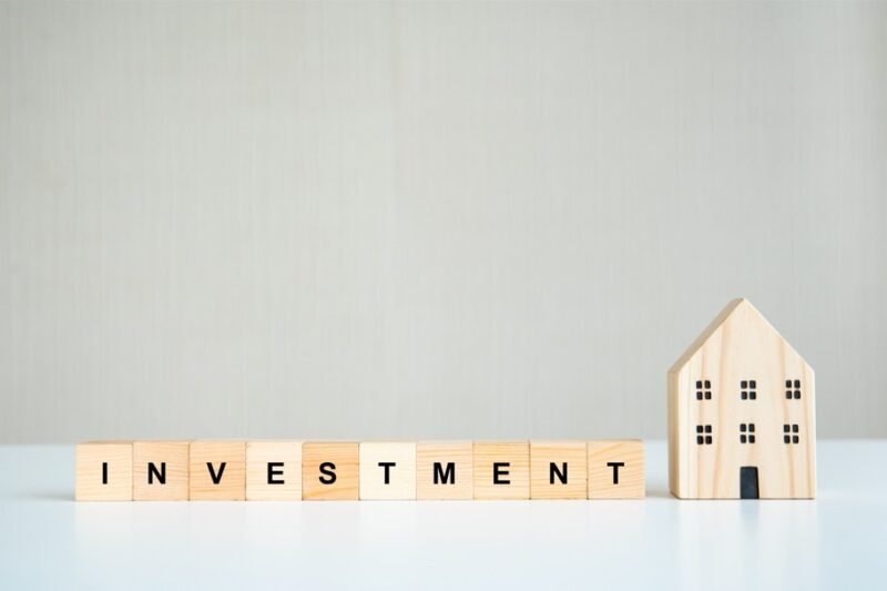 Des blocs de bois portant l'inscription "INVESTISSEMENT" sont disposés en rangée à côté d'une petite maquette de maison en bois sur une surface blanche à fond uni.