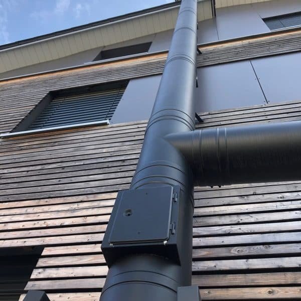 Tuyau de ventilation en métal noir fixé au mur extérieur en bois d'un bâtiment moderne, vu de dessous dans un ciel partiellement nuageux.