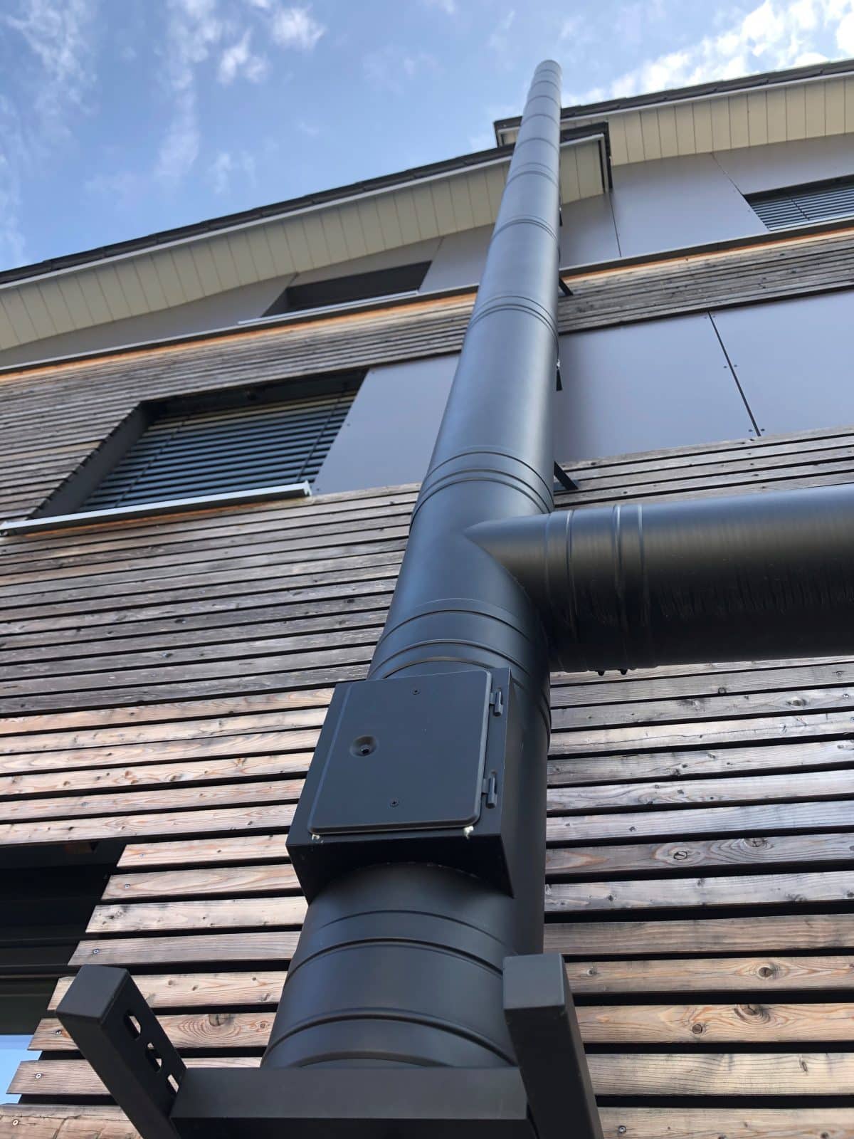 Tuyau de ventilation en métal noir fixé au mur extérieur en bois d'un bâtiment moderne, vu de dessous dans un ciel partiellement nuageux.