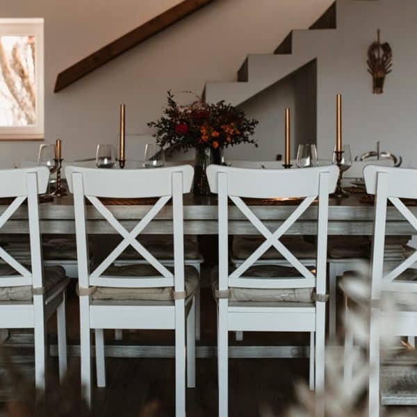 Salle à manger avec une table rectangulaire dressée pour un repas, avec des chaises blanches et des chandeliers. Un centre de table floral décore la table. Une petite fenêtre et un escalier sont visibles en arrière plan.