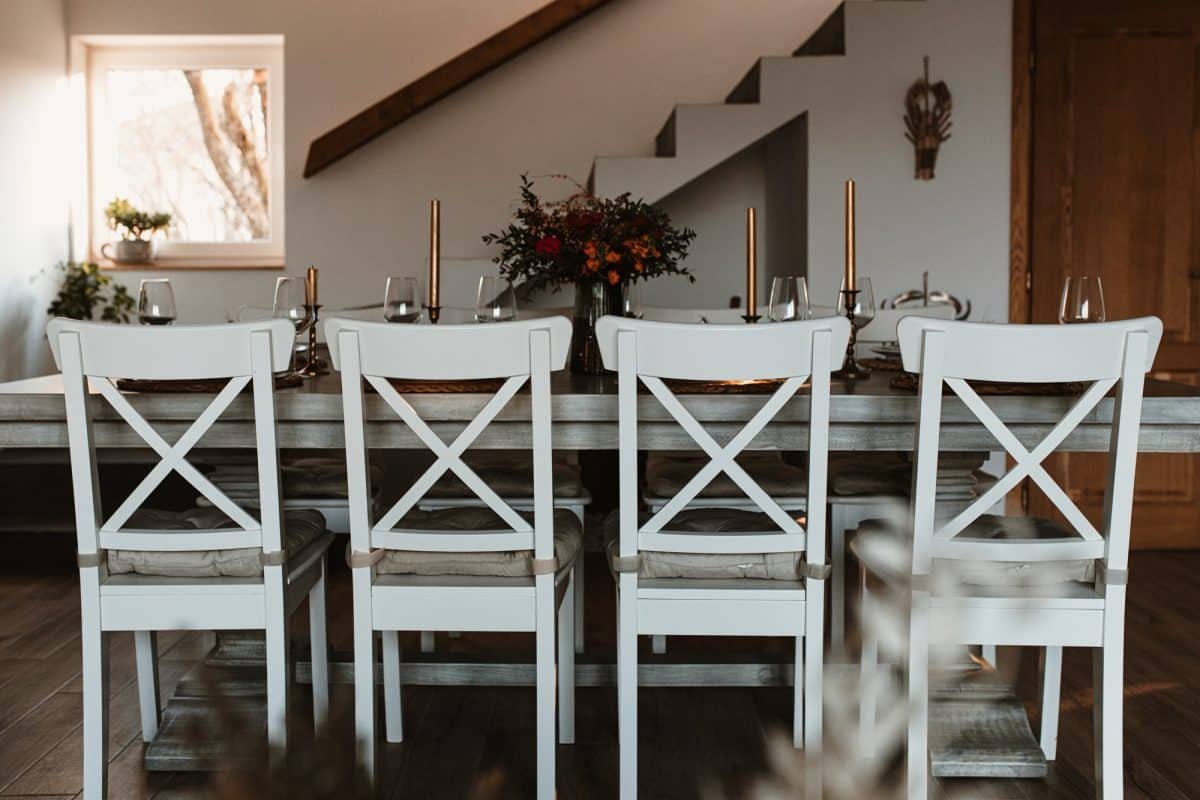Salle à manger avec une table rectangulaire dressée pour un repas, avec des chaises blanches et des chandeliers. Un centre de table floral décore la table. Une petite fenêtre et un escalier sont visibles en arrière plan.