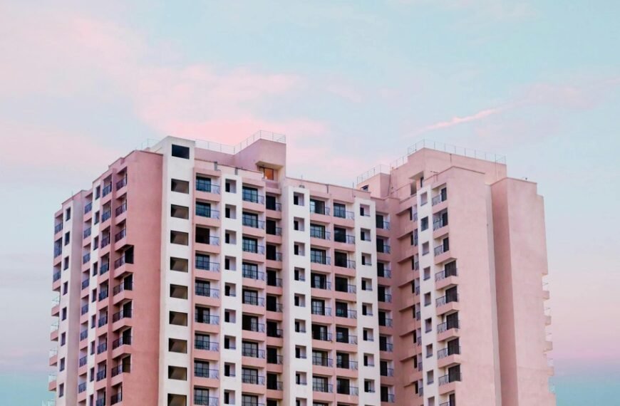 Un grand immeuble moderne aux multiples balcons et fenêtres se détache sur un ciel pastel aux teintes roses et bleues.