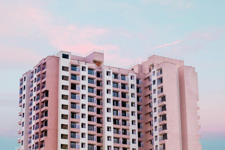 Un grand immeuble moderne aux multiples balcons et fenêtres se détache sur un ciel pastel aux teintes roses et bleues.