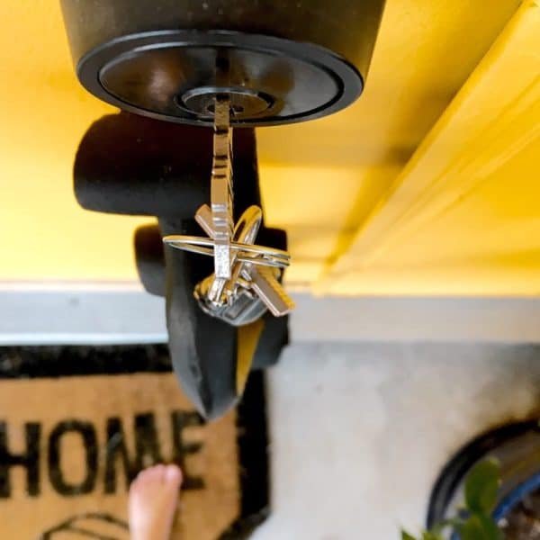 Un jeu de clés de maison inséré dans une serrure de porte au-dessus d'un paillasson sur lequel est écrit « MAISON », avec une partie d'une plante et un pied nu visibles à proximité.