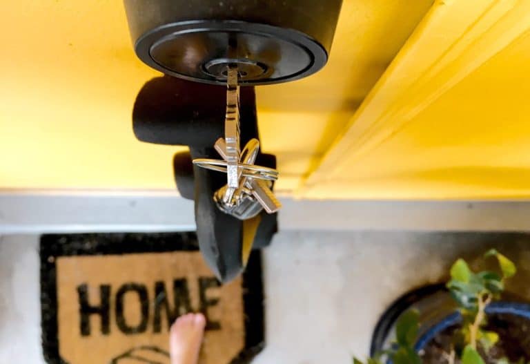 Un jeu de clés de maison inséré dans une serrure de porte au-dessus d'un paillasson sur lequel est écrit « MAISON », avec une partie d'une plante et un pied nu visibles à proximité.