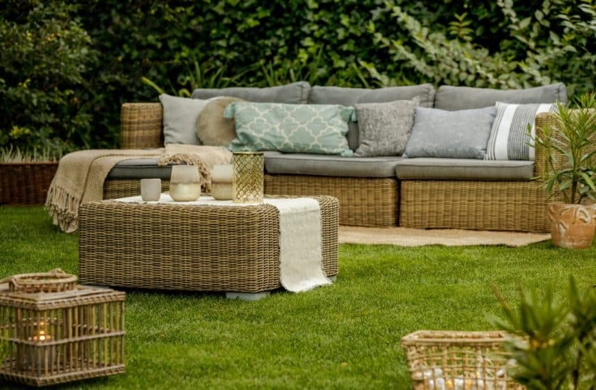 Ensemble de meubles d'extérieur en osier avec un canapé gris rembourré, des coussins à motifs et une table basse sur un espace gazonné. Une végétation luxuriante entoure le décor.