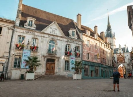 Bâtiment historique avec drapeaux et bannières, étiqueté « Hôtel de Ville », sur une place pavée. Une personne se tient debout au premier plan. Une tour d'horloge est visible à l'arrière plan.