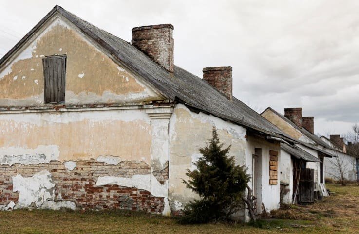 Vieux bâtiment usé par les intempéries, à la peinture écaillée, aux briques apparentes et aux multiples cheminées, entouré d'une pelouse envahie par la végétation, sous un ciel nuageux.