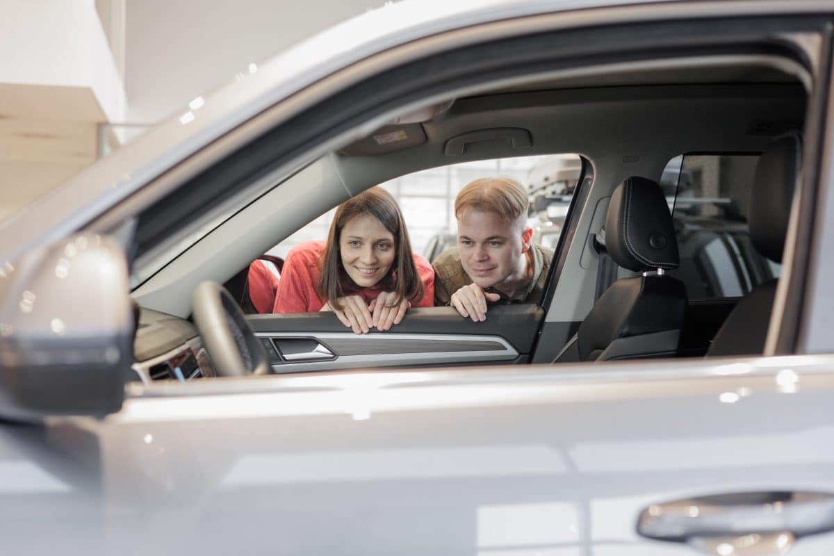 Deux personnes regardent l'intérieur d'une voiture garée à travers la vitre ouverte du côté conducteur dans une salle d'exposition.