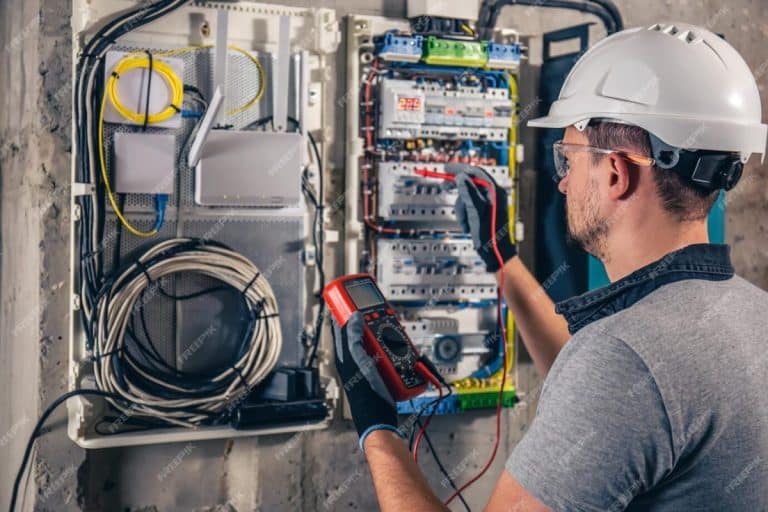 Un électricien portant un casque et des gants utilise un multimètre pour tester un panneau électrique dans une armoire murale, avec des câbles et des équipements organisés visibles.