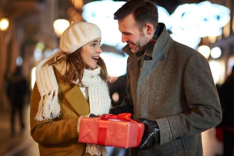Une femme portant une écharpe blanche et un béret reçoit joyeusement un cadeau emballé d'un homme portant un manteau gris dans une rue de la ville la nuit.