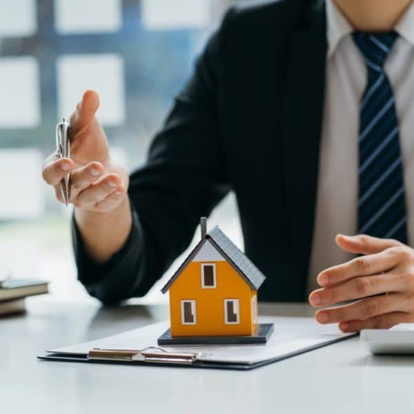 Une personne en costume discute d'investissement immobilier à un bureau avec une maison modèle, un presse papiers et une calculatrice.
